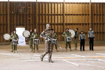 A markswoman holding a long rifle in front of drummers from a marching band and two football referees.