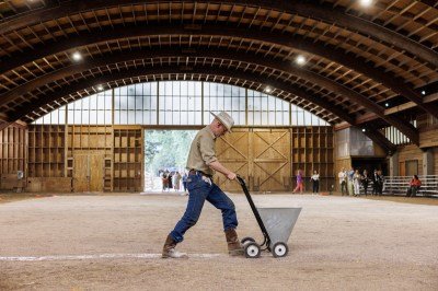 A man in cowboy clothes pushing a field-marking machine with a line of white chalk behind him.