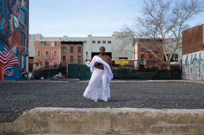 A Black woman with her white top blowing away to reveal her breasts standing in an empty lot.