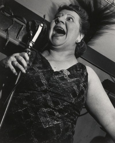 A black-and-white photograph of a woman singing into a microphone, causing her hair to spin above her head.