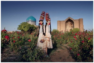 View of two women in traditional Kazakh dress standing in a garden with buildings behind. 