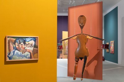 Installation view of a museum exhibition showing a painting of a boy at left and a sculpture resembling a human made of a violin and other found wooden objects. 