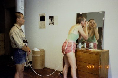 A man tied at the waist to a woman who does her make up using a mirror.
