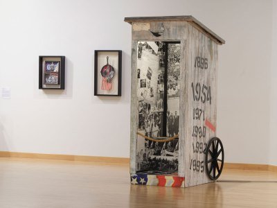 Two mixed-media artworks hang on a wall behind a sculpture that resembles an outhouse that has been decoupaged with photographs from the civil rights movement. 