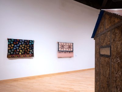 Two quilts hang on a wall. At left is a partial view of a sculpture resembling a shed. 
