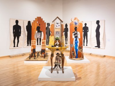 View of a multipart installation featuring various found objects on a table, two sculptures with human figures cut out, and nine wall-hung drawings with black silhouettes. 