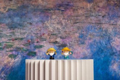 Two figurines of Claude Monet sit on a pedestal in front of a Monet 'water lily' painting.