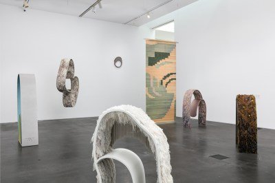 A gallery filled with sculptures, some of which are freestanding in the center. They feature ridged forms covered in dyed threads.